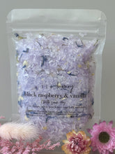Black raspberry and vanilla bath soak in a clear resealable pouch with pastel purple crystals and dried botanicals.