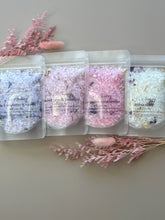Pastel bath soak pouches in assorted scents displayed side by side, including blackberry and vanilla, sweet pea and jasmine, champagne strawberries and Miss Dior, styled with pink dried florals